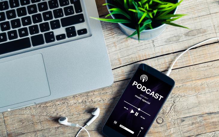 An iPhone playing a podcast with a mac book and plant pot next to it