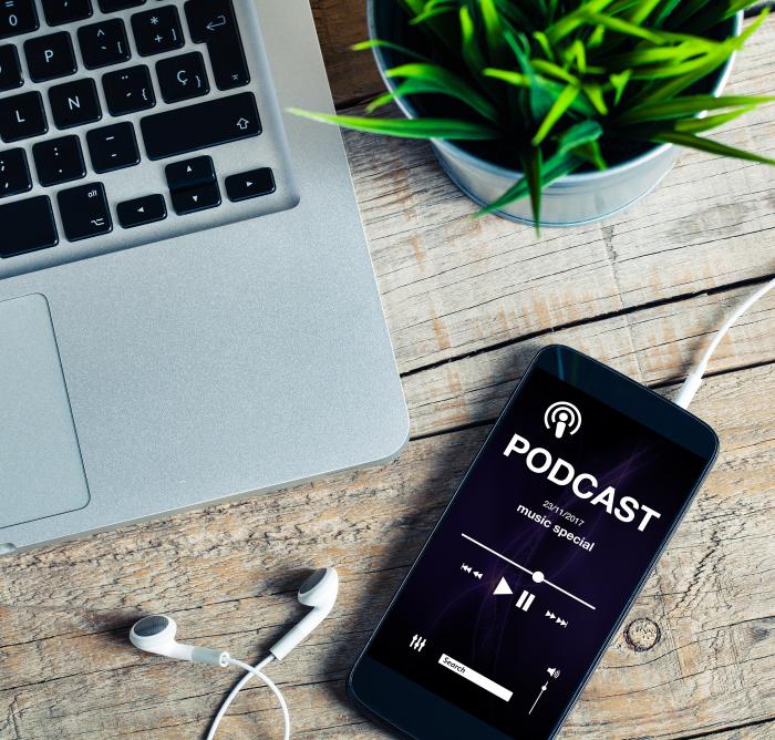 An iPhone playing a podcast with a mac book and plant pot next to it